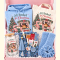 A curated Christmas gift box set with matching printed items, including a light blue hoodie, a cream tote bag, a glass mason jar, blue plush socks, a blue scrunchie, and small gift items, all arranged in a pink box with white filler. The printed designs feature a Christmas fireplace scene and the text 'All Booked for Christmas'.