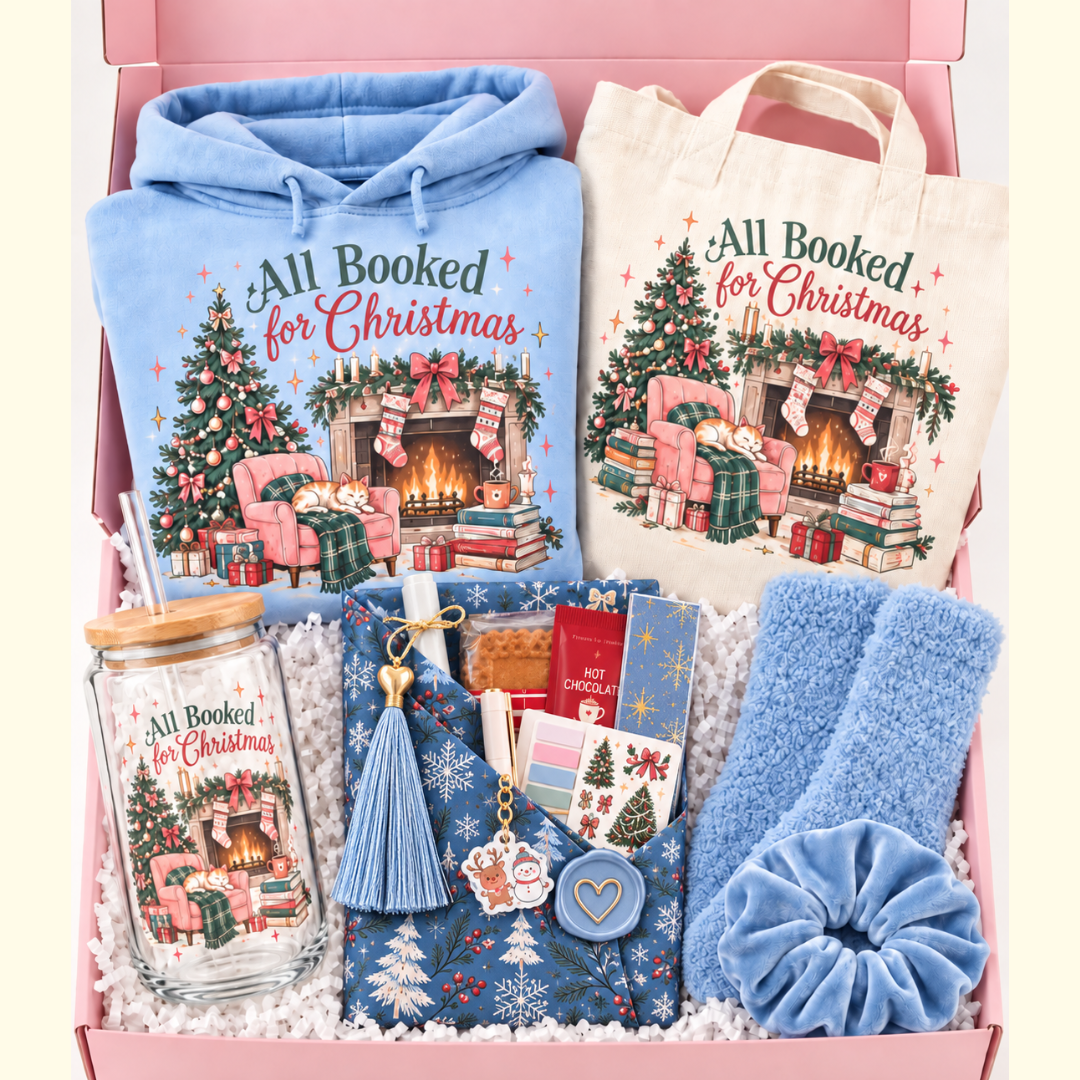 A curated Christmas gift box set with matching printed items, including a light blue hoodie, a cream tote bag, a glass mason jar, blue plush socks, a blue scrunchie, and small gift items, all arranged in a pink box with white filler. The printed designs feature a Christmas fireplace scene and the text 'All Booked for Christmas'.