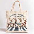 A cream canvas tote bag with a festive Christmas graphic featuring four deer in colorful knit sweaters walking across a snowy crosswalk, with the text 'DASHING THROUGH THE SNOW' printed above them.