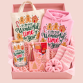 A pink gift box filled with holiday-themed items, including a pink hoodie and tote bag printed with gingerbread illustrations and the phrase 'it's the most Wonderful time of the Year,' a matching pink scarf, striped socks, a glass mason jar, and small gift items.