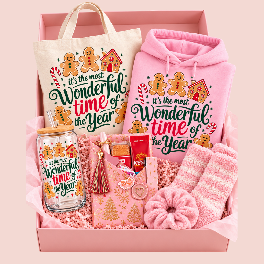 A pink gift box filled with holiday-themed items, including a pink hoodie and tote bag printed with gingerbread illustrations and the phrase 'it's the most Wonderful time of the Year,' a matching pink scarf, striped socks, a glass mason jar, and small gift items.